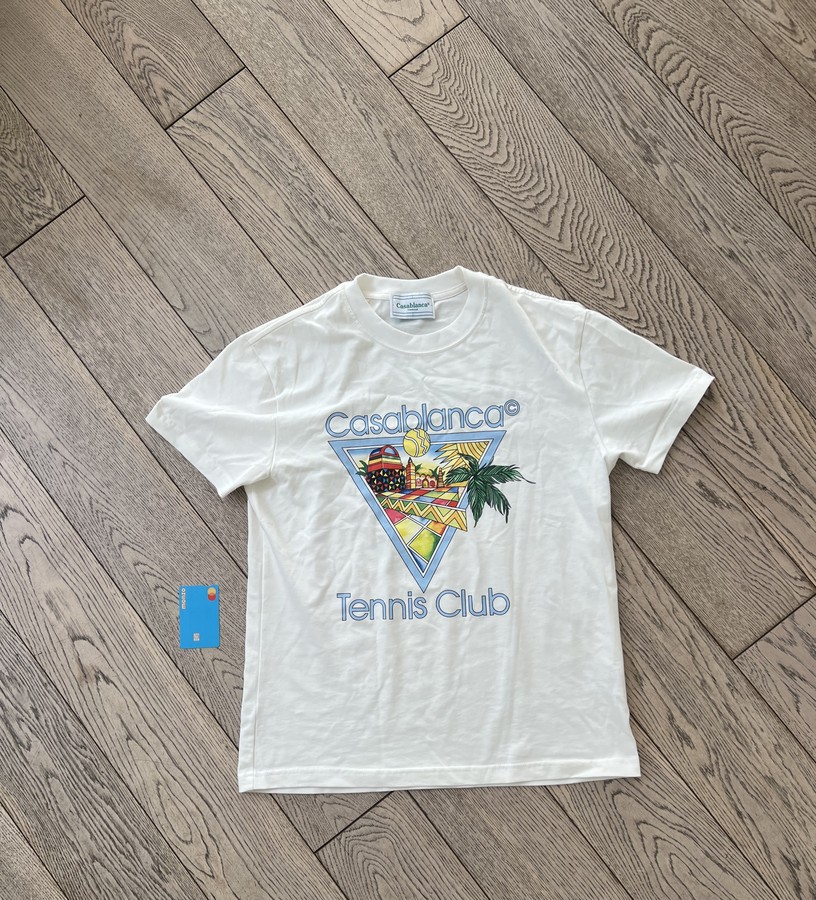 A cream Casablanca t-shirt laid flat on a wooden floor with a bank card placed beside it, ready to be photographed